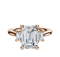 The 18K Rose Gold 3-Stone Emerald Cut Side Stones 0.125TCW Engagement Ring features a central emerald-cut diamond with two smaller rectangular diamonds on each side, shown on a plain white background.