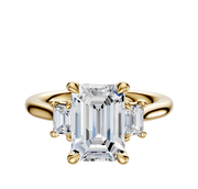 The 14K Yellow Gold 3-Stone Emerald Cut Side Stones 0.125TCW Engagement Ring features a center emerald-cut diamond with two smaller rectangular diamonds on each side, all set in a classic four-prong setting.