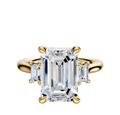 The 18K Yellow Gold 3-Stone Emerald Cut Side Stones 0.25TCW Engagement Ring features a central emerald-cut diamond with two smaller rectangular diamonds on each side, set in classic prongs against a white backdrop.