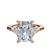 The 18K Rose Gold 3-Stone Trillion Side Stones 0.125TCW Engagement Ring features a radiant-cut center diamond and two smaller trillion side stones, elegantly set against a white background.