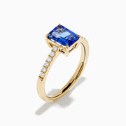 The Nahla 14K Yellow Gold Tanzanite and Diamond Ring showcases a rectangular blue tanzanite center stone, accented by small round white diamonds along the upper band on a plain white background—timeless elegance in every detail.