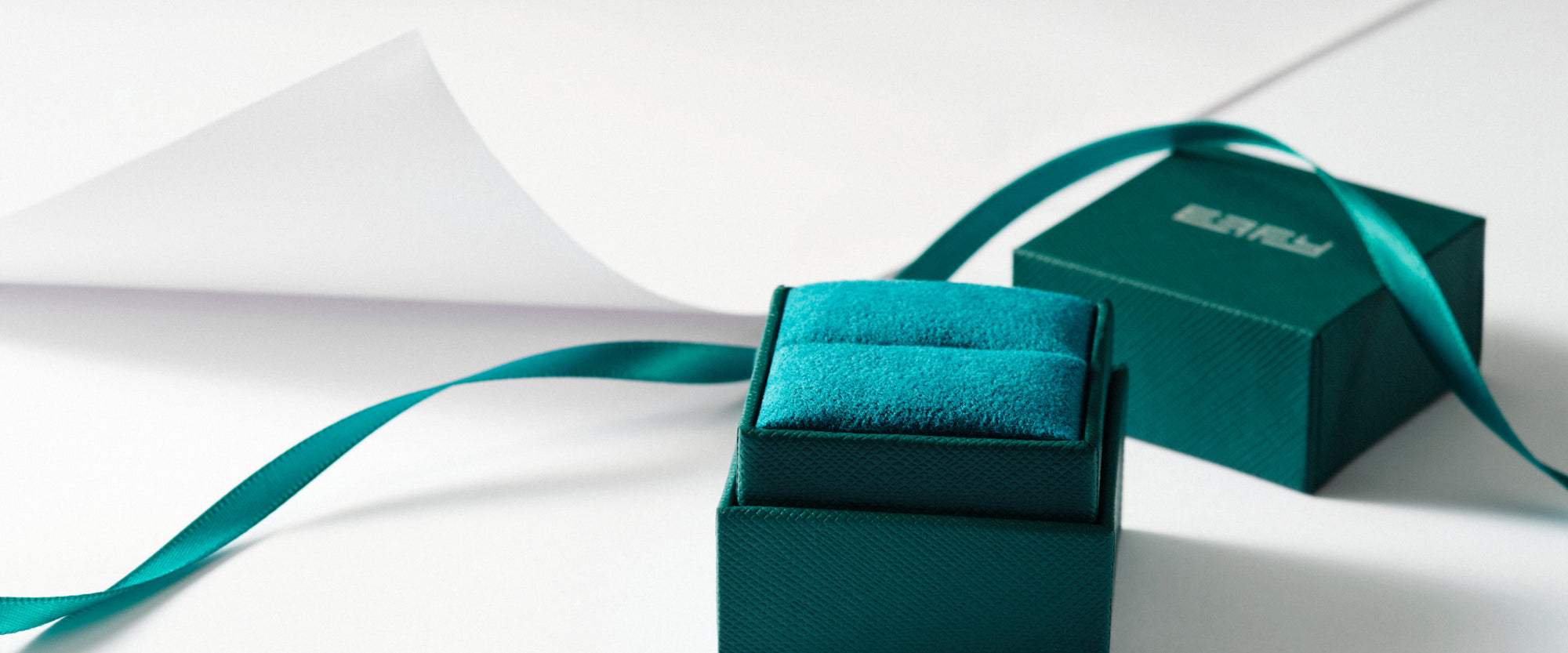 A teal-colored jewelry box with a matching ribbon sits open on a white surface, revealing a soft interior. Another similar box and ribbon are in the background.