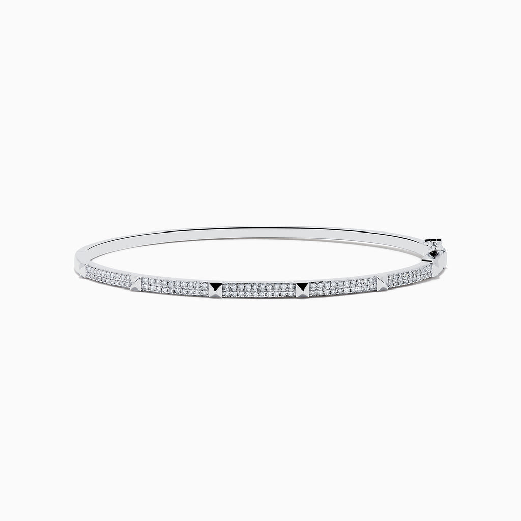 The 14K White Gold Diamond Spike Bangle showcases alternating pyramid studs and rows of sparkling diamonds on its sleek silver surface, set against a plain white background.