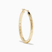 The 14K Yellow Gold Crystal Cut Tube Oval Hoops feature a textured, engraved floral pattern and delicate crystal cut detailing, shown against a plain white background.