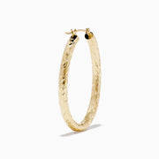 The 14K Yellow Gold Crystal Cut Tube Oval Hoops feature a textured, engraved floral pattern and delicate crystal cut detailing, shown against a plain white background.