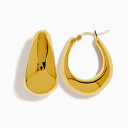 The 14K Yellow Gold Hollow Hoops feature a shiny, abstract shape with smooth, curved surfaces, reminiscent of classic hoops, and are displayed against a white background.