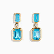Elegant and symmetrical, the 14K Yellow Gold Blue Topaz and Diamond Drop Earrings display rectangular blue topaz stones with smaller gems above, separated by a sparkling diamond drop, set against a white background.