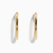 The 14K Yellow Gold Hollow Tube Hoops are sleek, polished earrings shown side by side against a white background, featuring classic yellow gold in a timeless hoop design with clasps at the top.