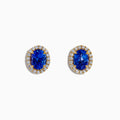 The Nahla 14K Yellow Gold Tanzanite and Diamond Stud Earrings feature oval blue tanzanites each encircled by a halo of diamonds, beautifully displayed on a white background.
