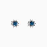 Bella Bleu 14K White Gold Blue and White Diamond Earrings, 1.20 TCW, feature a central round blue stone encircled by sparkling white diamonds in a flower-inspired design, displayed on a white background.