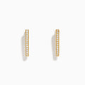 A pair of D'Oro 14K Yellow Gold Diamond Triangle Huggie Earrings are shown on a plain white background, each featuring a row of sparkling diamonds.