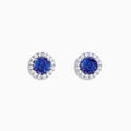 The Nahla 14K White Gold Tanzanite and Diamond Stud Earrings feature round deep blue tanzanite centers, each surrounded by a halo of sparkling white diamonds, beautifully set in 14K white gold against a crisp white background.