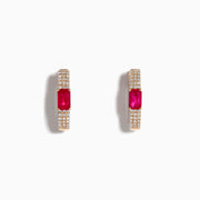 The Ruby Royale 14K Rose Gold Ruby and Diamond Huggie Earrings feature rectangular red rubies flanked by rows of sparkling white diamonds, set in rose gold and displayed against a white background.