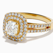 The 18K Yellow Gold Diamond Double Halo Ring showcases a cushion-cut center stone, surrounded by a double halo of round diamonds and additional diamonds set along the split shank band.