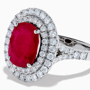 The 18K White Gold Ruby and Diamond Double Halo Ring features an oval ruby encircled by two rows of round white diamonds, set in a sleek 18K white gold band and displayed on a white background.