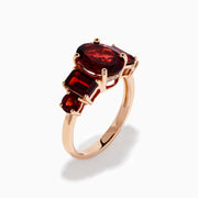 The 14K Rose Gold Multi Shape Garnet and Diamond Hidden Halo Ring showcases a central oval deep red garnet, accented by smaller rectangular and oval garnets on each side, all set in polished rose gold.