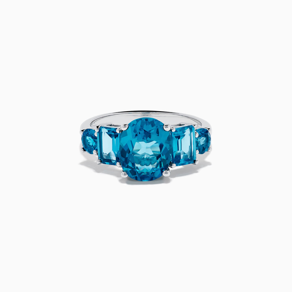 The 14K White Gold Multi Shape Blue Topaz and Diamond Hidden Halo Ring showcases a bold oval blue topaz center, accented by rectangular and round blue stones on each side, all set in a polished band with a subtle diamond hidden halo.