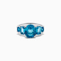 The 14K White Gold Multi Shape Blue Topaz and Diamond Hidden Halo Ring showcases a bold oval blue topaz center, accented by rectangular and round blue stones on each side, all set in a polished band with a subtle diamond hidden halo.