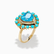 The 14K Yellow Gold Blue Topaz, Turquoise and Diamond Heart Cocktail Ring features a striking heart-shaped blue topaz center, surrounded by a halo of diamonds and an outer border of vibrant round turquoise stones.