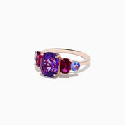 The 14K Rose Gold Amethyst, Rhodolite, and Tanzanite Ring features a central oval amethyst accented by smaller red rhodolite and blue tanzanite gems on each side, displayed on a white background.