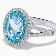 The Aquarius 14K White Gold Aquamarine, Paraiba, and Diamond Ring features an oval aquamarine center stone encircled by two halos of sparkling blue Paraiba and clear diamonds, displayed on a plain white background.