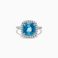 The 14K White Gold Blue Zircon and Diamond Ring showcases a large cushion-cut blue zircon center, surrounded by a halo of small round diamonds on the setting and band, set against a white background.