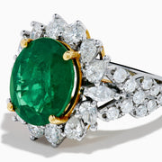 The 18K Two-Tone Gold Emerald, Yellow and White Diamond Ring features a striking oval green emerald surrounded by white diamonds, set in a gold and silver band with gold prongs, shown against a plain white background.