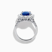 The 18K White Gold Blue Sapphire and Diamond Halo Ring features a large oval blue sapphire surrounded by a halo of round diamonds, showcased on a white background.