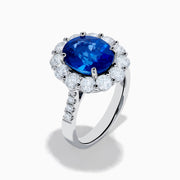 The 18K White Gold Blue Sapphire and Diamond Halo Ring features a large oval blue sapphire surrounded by a halo of round diamonds, showcased on a white background.