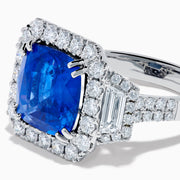 The Royale Bleu 18K White Gold Blue Sapphire and Diamond Halo Ring showcases a cushion-cut blue sapphire center with a double diamond halo and diamond side accents, elegantly set against a white background.
