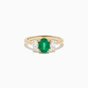 The 14K Yellow Gold Emerald and Diamond Three-Stone Ring showcases an oval green emerald center with two oval white diamonds on each side, all set in 14K yellow gold and displayed against a white background.