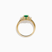 The 14K Yellow Gold Emerald and Diamond Three-Stone Ring showcases an oval green emerald center with two oval white diamonds on each side, all set in 14K yellow gold and displayed against a white background.