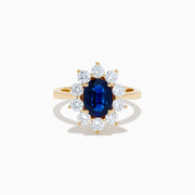The 14K Yellow Gold Natural Blue Sapphire and Diamond Lady Di Ring features an oval sapphire center, haloed by round white diamonds, shown on a white background.