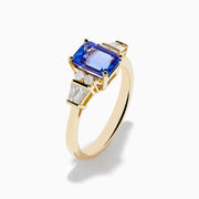 The Nahla 14K Yellow Gold Tanzanite and Diamond Ring showcases a rectangular blue tanzanite center, with two round diamonds and two baguette-cut white stones on each side, displayed on a white background.