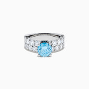 The Lab Grown Blue Diamond Round Brilliant Solitaire Ring in 14K White Gold features a striking round blue diamond center, accented by two rows of sparkling clear diamonds along the band, shown on a white background.