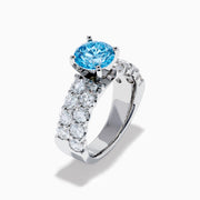 The Lab Grown Blue Diamond Round Brilliant Solitaire Ring in 14K White Gold features a striking round blue diamond center, accented by two rows of sparkling clear diamonds along the band, shown on a white background.