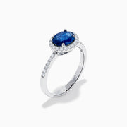 The 14K White Gold East West Oval Blue Sapphire and Diamond Halo Ring showcases an oval blue sapphire encircled by a diamond halo, with extra diamonds accenting the band for added brilliance.