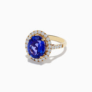 The Nahla Siri 14K Yellow Gold Tanzanite and Diamond Ring features a large oval blue tanzanite center stone encircled by a halo of small round white diamonds, displayed on a plain white background.