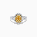 The Canare 18K Two Tone Gold White and Yellow Oval Diamond Split Shank Ring showcases an oval yellow diamond center with a double halo of white diamonds and a diamond-accented split shank, displayed on a plain white background.