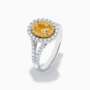 The Canare 18K Two Tone Gold White and Yellow Oval Diamond Split Shank Ring showcases an oval yellow diamond center with a double halo of white diamonds and a diamond-accented split shank, displayed on a plain white background.