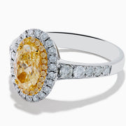 The Canare 18K Two Tone Gold White and Yellow Oval Diamond Ring features a large yellow diamond at the center, a halo of small round white diamonds, and additional diamonds set along the elegant band.