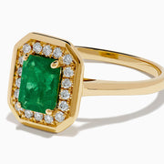 The Brasilica 14K Yellow Gold Emerald and Diamond Ring showcases a rectangular green emerald center, surrounded by a halo of sparkling round white diamonds, all set in yellow gold and displayed on a white background.