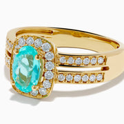 The 18K Yellow Gold Paraiba and Diamond Halo Ring showcases a central oval Paraiba turquoise, surrounded by a diamond halo and additional diamonds on the split shank band, all set in yellow gold and shown against a white background.
