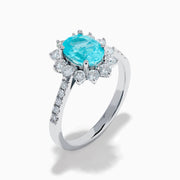 The 14K White Gold Paraiba and Diamond Halo Ring showcases an oval-shaped Paraiba gemstone at the center, encircled by a sparkling diamond halo with additional diamonds adorning the band.