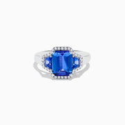 The Nahla 14K White Gold 3 Stone Tanzanite and Diamond Ring features an emerald-cut tanzanite center flanked by sparkling diamonds, all set in a geometric design and displayed on a white background.