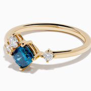 The 14K Yellow Gold Blue and White Diamond Ring showcases a cushion-cut blue stone center with two round white diamonds on each side, set against a white background.