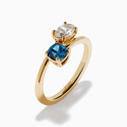 The Bella Bleu 14K Yellow Gold Blue and Light Brown Bypass Diamond Ring showcases an oval white diamond and a cushion-cut deep blue gemstone set side by side on a sleek gold band.