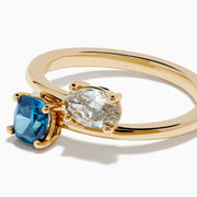 The Bella Bleu 14K Yellow Gold Blue and Light Brown Bypass Diamond Ring showcases an oval white diamond and a cushion-cut deep blue gemstone set side by side on a sleek gold band.