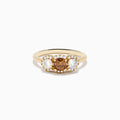 The Espresso 14K Yellow Gold Brown Diamond 3-Stone Ring features a central brown diamond, two oval white stones, and sparkling halo accents for a stunning look.