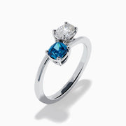 The Bella Bleu 14K White Gold Bypass Ring showcases a blue stone and a light brown diamond set side by side in prongs, displayed against a plain white background.